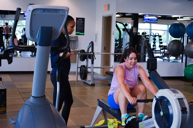 Smiling participant on a rowing machine alongside a trainer at a group fitness class at the fitness centre at the Paramount Fine Foods Centre